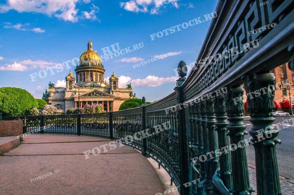 Фотография показывает величественный Исаакиевский собор в Санкт-Петербурге, снятый с уровня чугунного ограждения одного из мостов. Чёткое голубое небо и лёгкие облака создают яркий контраст с золотым куполом собора. Такой снимок отлично подходит для путеводителей по городу, исторических и архитектурных обзоров, а также может использоваться в качестве учебного материала или постера.